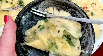 A hand holds a dark ceramic bowl with thinly sliced pineapple, garnished with fresh herbs. In the background, more pineapple slices with herbs and red chilli are arranged on a white plate. A small fork rests in the bowl.