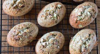 Oval-shaped baked goods topped with chopped nuts are cooling on a black wire rack placed on a wooden surface. The golden-brown pastries appear fluffy and freshly baked.