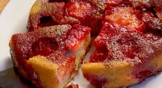 A close-up of a plum upside-down cake on a white plate, with a slice being lifted out. The cake has caramelised plums on top and a golden, moist-looking base. The background is a wooden surface.