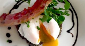 A poached egg with a runny yolk sits on a dark patty, garnished with microgreens and chives, topped with a crispy rasher of bacon, and surrounded by a swirl of dark sauce on a white plate.