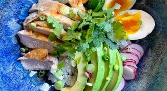 A blue bowl filled with rice, sliced grilled chicken, avocado, soft-boiled egg, cucumber, radish, coriander, and spring onions, arranged neatly for a colourful and fresh meal.