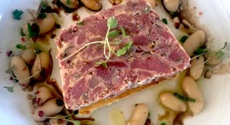 A rectangular slice of terrine garnished with microgreens sits in the centre of a plate, surrounded by white beans, chopped herbs, and a drizzle of sauce.