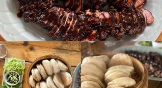 A platter of sliced, glazed pork fillet, a bamboo steamer filled with fluffy bao buns, and plates of spring onions, cucumber, and hoisin sauce arranged on a wooden table.