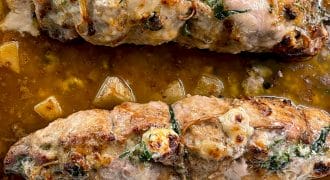 Two stuffed pork fillets tied with string are baked in a brown sauce with visible herbs and chunks of vegetables in a rectangular roasting tin.