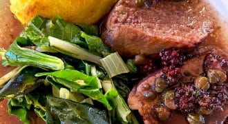 A plate with two slices of beef topped with capers and berries in sauce, sautéed leafy greens, and a round serving of cornbread or a baked yellow side.