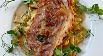 A white plate with pasta topped with a slice of prosciutto-wrapped meat, creamy sauce, and garnished with fresh pea shoots.