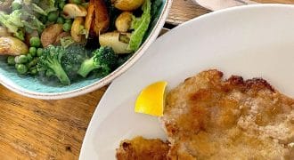 A white plate with two breaded cutlets and a lemon wedge, beside a bowl of roast potatoes, broccoli, peas, and greens on a wooden table.