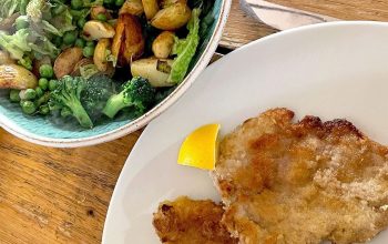 A white plate with two breaded cutlets and a lemon wedge, beside a bowl of roast potatoes, broccoli, peas, and greens on a wooden table.