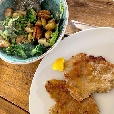 A white plate with two breaded cutlets and a lemon wedge, beside a bowl of roast potatoes, broccoli, peas, and greens on a wooden table.