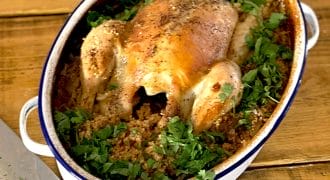 A whole roast chicken sits atop a bed of seasoned grains and herbs in an oval casserole dish, garnished with chopped parsley, on a wooden table beside a carving knife.