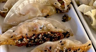 A close-up of pan-fried dumplings topped with white and black sesame seeds, arranged in a white rectangular dish alongside steamed dumplings.
