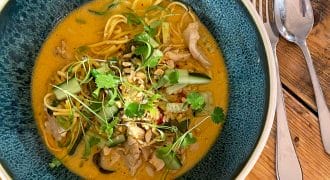 A bowl of noodle soup with sliced vegetables, herbs, and chicken in a creamy, orange broth, served on a rustic wooden table with a spoon and fork beside the bowl.