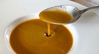 A spoon pours thick brown gravy into a shallow white bowl, with some gravy already pooled at the bottom. The scene is set on a wooden table with a blue and white cup in the background.