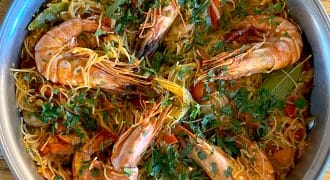 A metal pan filled with cooked noodles, large prawns, chopped tomatoes, and garnished with fresh herbs, sits on a wooden table.