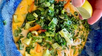 A hand squeezes a lime wedge over a bowl of colourful curry soup with noodles, leafy greens, carrots, and chopped fresh herbs in a blue dish.