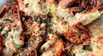 Grilled prawns with heads on, served on a white platter, topped with a creamy herb and lemon sauce, sprinkled with fresh herbs and red pepper flakes.