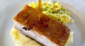 A thick slice of crispy roast pork belly sits on mashed potatoes, with gravy being poured over it. A side of cooked cabbage is also on the white plate.