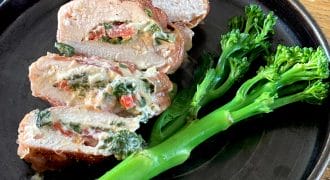 Sliced stuffed chicken breast filled with spinach, cheese, and red peppers, served on a dark plate with steamed tenderstem broccoli on the side, placed on a wooden table.