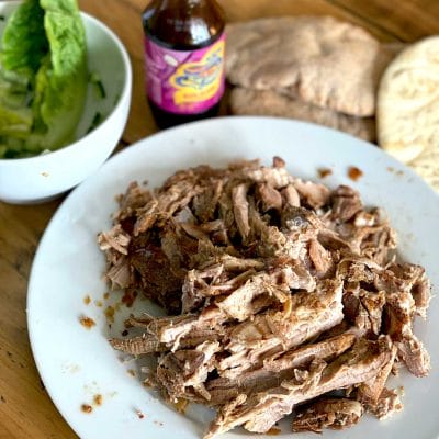 Shredded cooked meat on a white plate, with pitta bread, a bottle of sauce, and a bowl of lettuce and cucumber on a wooden table.