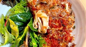 A white plate with a serving of aubergine Parmesan topped with tomato sauce and melted cheese, next to a side of mixed leafy green salad.