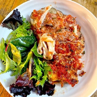 A white plate with a serving of aubergine Parmesan topped with tomato sauce and melted cheese, next to a side of mixed leafy green salad.