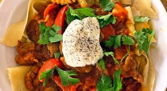 A white plate of wide pasta topped with a tomato-based meat stew, red peppers, fresh parsley, and a dollop of soured cream sprinkled with black pepper, served on a wooden table.