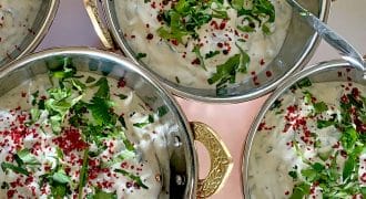 Four metal bowls with ornate handles are filled with creamy raita, garnished with chopped fresh herbs and red spices. Spoons rest in each bowl, and the bowls are arranged closely together on a light surface.