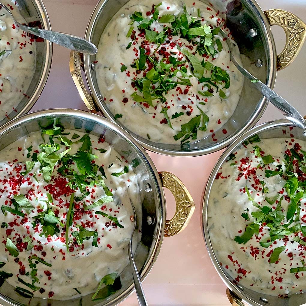 Four metal bowls with ornate handles are filled with creamy raita, garnished with chopped fresh herbs and red spices. Spoons rest in each bowl, and the bowls are arranged closely together on a light surface.