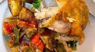 A plate with a roasted chicken leg, roast potatoes, and a colourful mix of cooked vegetables, including tomatoes, peppers, and aubergine, with a fork and knife on the side.