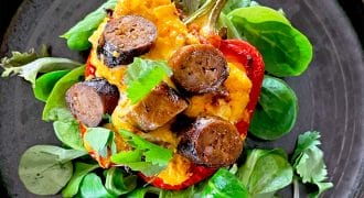 A halved red pepper filled with mashed potato and sliced sausage, garnished with fresh herbs and parsley, served on a dark plate with leafy greens underneath.