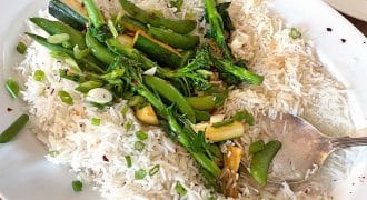 A white oval plate with steamed white rice topped with green vegetables, including sprouting broccoli, mangetout, and courgette, garnished with sliced spring onions and red chilli flakes. A serving spoon rests on the plate.