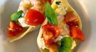 Two pasta shells filled with creamy cheese, prawns, chopped tomatoes, and fresh basil leaves, served on a white plate.