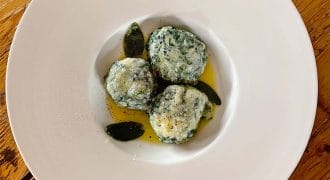 Three spinach and ricotta dumplings served on a white plate with sage leaves and melted butter, placed on a rustic wooden table.