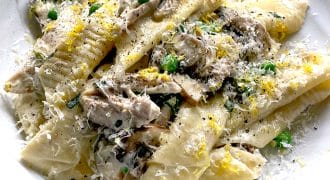 A plate of wide, homemade pasta with creamy mushroom sauce, topped with grated cheese, chopped herbs, and lemon zest, served on a white dish.