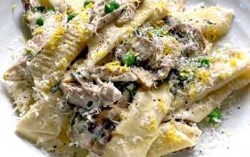 A plate of wide, homemade pasta with creamy mushroom sauce, topped with grated cheese, chopped herbs, and lemon zest, served on a white dish.