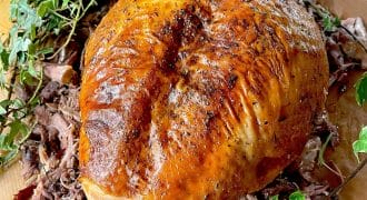 A golden-brown, roasted turkey breast is surrounded by shredded turkey meat and garnished with fresh green herbs on a wooden surface.