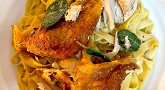 A plate of fettuccine pasta topped with roasted chicken pieces and crispy sage leaves, served on a white dish.
