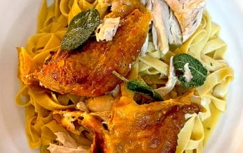 A plate of fettuccine pasta topped with roasted chicken pieces and crispy sage leaves, served on a white dish.