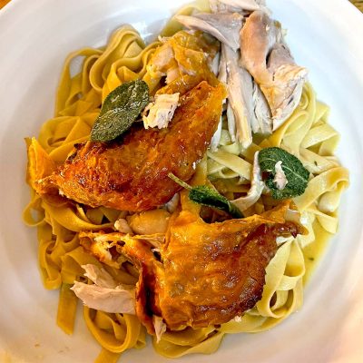 A plate of fettuccine pasta topped with roasted chicken pieces and crispy sage leaves, served on a white dish.