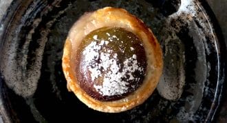 A round pastry with a glossy, dark filling dusted with icing sugar sits on a rustic, dark ceramic plate with a swirl of white sauce.