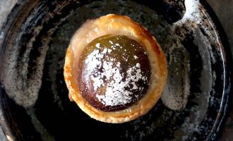 A round pastry with a glossy, dark filling dusted with icing sugar sits on a rustic, dark ceramic plate with a swirl of white sauce.