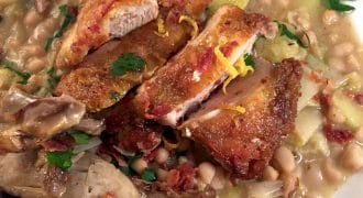 A close-up of a plate with crispy sliced chicken on top of a hearty white bean stew, garnished with fresh parsley and lemon zest. The stew contains beans, leeks, and shredded meat.