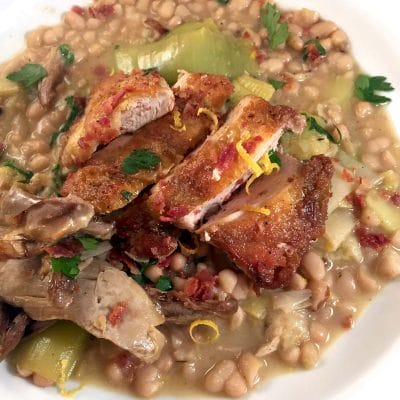 A close-up of a plate with crispy sliced chicken on top of a hearty white bean stew, garnished with fresh parsley and lemon zest. The stew contains beans, leeks, and shredded meat.