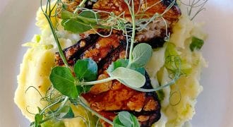Grilled tempeh served on a bed of mashed potato, garnished with fresh pea shoots and microgreens, presented on a white plate.