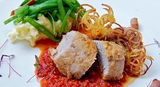 Two thick slices of cooked meat sit on a bed of red tomato sauce, served with mashed potato, green beans, crispy shoestring chips, and garnished with microgreens on a white plate.