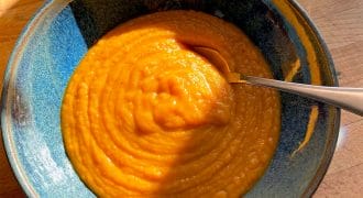 A blue ceramic bowl filled with thick, orange soup sits on a wooden table, with a spoon inside. Sunlight and shadows fall across the bowl, and a glass of water is visible in the background.