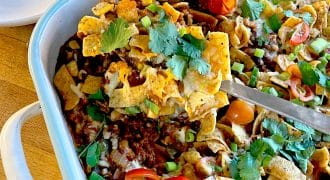 A casserole dish filled with a layered Frito pie topped with corn chips, melted cheese, minced beef, tomatoes, spring onions, and coriander. A serving is being lifted out with a spatula. A stack of white plates is nearby.