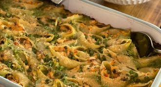 A large white baking dish filled with stuffed pasta shells topped with melted cheese and green herbs, with a serving spoon inside. In the background, there is a bowl of salad and a bottle of olive oil.