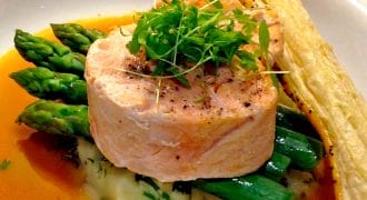 A gourmet dish featuring a round piece of salmon atop mashed potato with green asparagus spears, garnished with microgreens, served with a brown sauce and a puff pastry stick on the side.