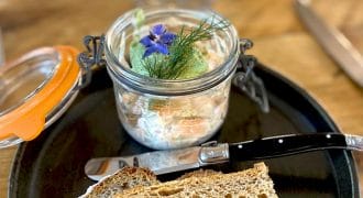 A black plate with two slices of rustic bread, a knife, and an open glass jar containing a creamy spread garnished with fresh herbs and edible flowers. The background shows a wooden table and a blurred wine glass.